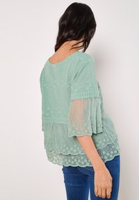 Mintgrünes Blusenshirt aus transparentem, besticktem Stoff mit kurzen Glockenärmeln und gewelltem Saum. Zarte Blumenmuster schmücken das Design.