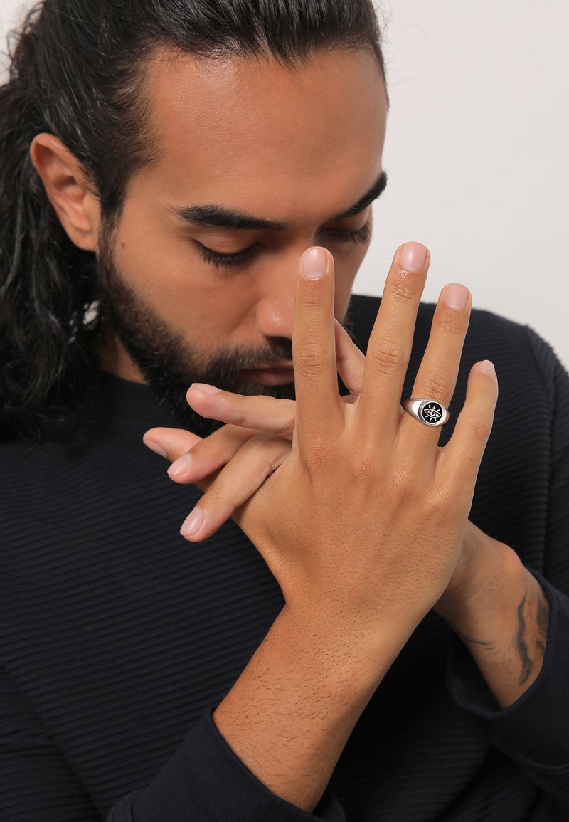 Silberner Ring mit schwarzem Auge-Design, an einer Hand mit hellbrauner Haut getragen. Der Hintergrund zeigt ein strukturiertes navyblaues Hemd. Die Hände sind nachdenklich positioniert.