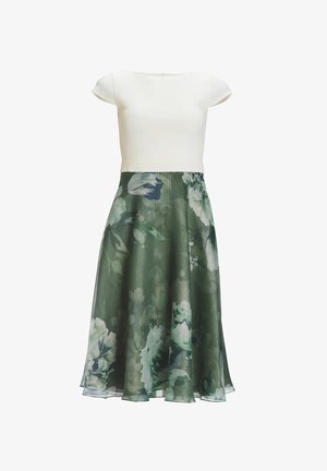 Een getailleerde witte top met korte mouwen gaat over in een vloeiende groene rok met bloemmotieven in blauwe en witte tinten, van gestructureerde stof.