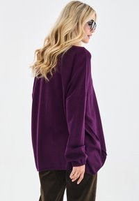 Blondynka w oversize fioletowym swetrze i ciemnych spodniach, stojąca bokiem, w okularach przeciwsłonecznych i z falującymi włosami na tle białego tła.