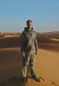 Hoodie w kamuflażu z naszywką na froncie, w komplecie z lekkimi spodniami cargo w kolorze oliwkowym oraz czarnymi butami, na tle piaszczystych wydm.