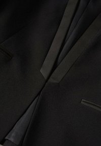 Černý blazer s hladkým materiálem, ostrými klopami a jemnými kapsami, který zdůrazňuje přizpůsobený a strukturovaný design.
