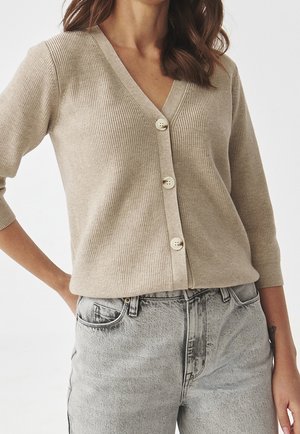 Beżowy kardigan z prążkowanej dzianiny z głębokim dekoltem w kształcie V, rękawami o długości trzech ćwierć i pięcioma jasnymi guzikami. Połączony z jasnoszarymi jeansami z wysokim stanem.