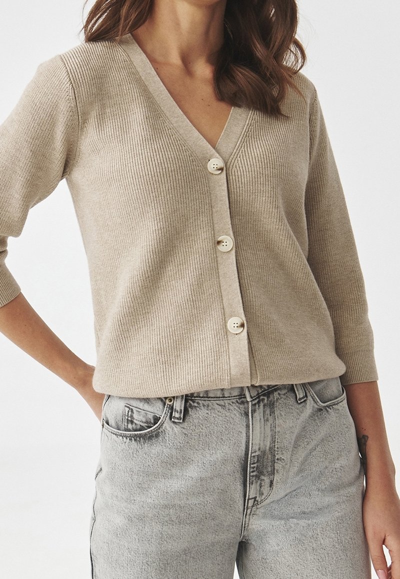 Beżowy kardigan z prążkowanej dzianiny z głębokim dekoltem w kształcie V, rękawami o długości trzech ćwierć i pięcioma jasnymi guzikami. Połączony z jasnoszarymi jeansami z wysokim stanem.