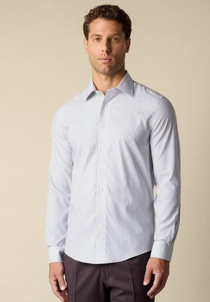 Mężczyzna ubrany w białą koszulę z długim rękawem, ozdobioną cienkimi pionowymi niebieskimi paskami, oraz ciemne spodnie, stojący na tle jednolitego beżowego tła.