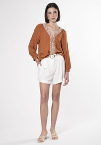 Blusa color ruggine con punti in rilievo, dettaglio floreale sul colletto e maniche lunghe; abbinata a pantaloni corti di lino bianchi e sandali bianchi con cinturini.