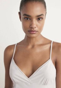Femme à la peau lisse et foncée, les cheveux tirés en arrière, portant un débardeur blanc à fines bretelles, de face sur un fond uni.