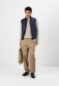 Marineblå puffervest over en beige strukturert genser, paret med beige bukser og sorte sko, holder en kremfarget totebag.