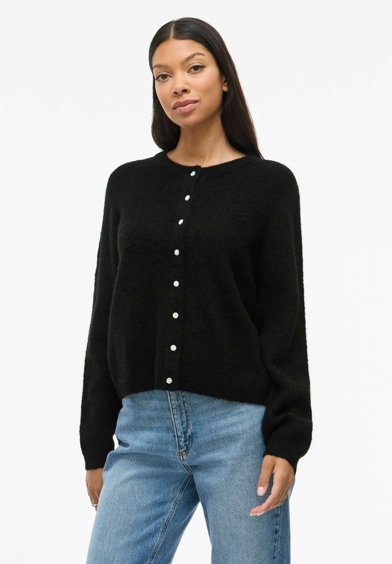 Cardigan en tricot noir avec un col rond, fermeture à boutons à l'avant et manches longues. Texture douce, coupe décontractée, associé à un jean bleu.