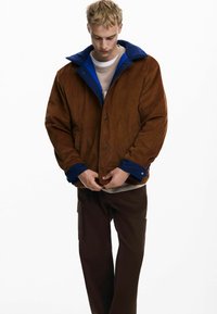 Braune Cordjacke mit blauen Kragen, ausgestattet mit Druckknöpfen, getragen über einem beigen Hemd und kombiniert mit braunen Cargo-Hosen.