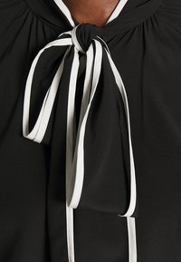 Schwarzes Oberteil mit gerafftem Ausschnitt und gebundenem Schleifendetail, das kontrastierende weiße Streifen aufweist und einen schichtigen Textureneffekt erzeugt.