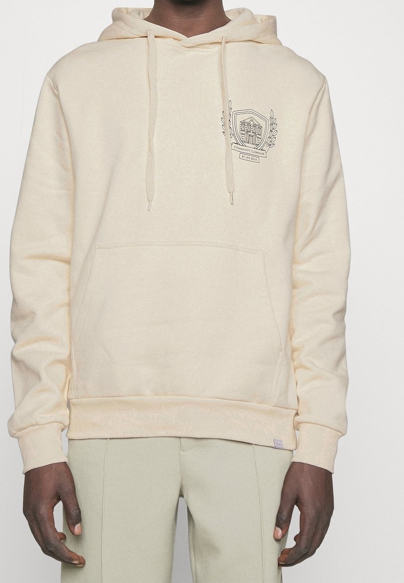 Sudadera con capucha beige con bolsillo frontal, capucha con cordón ajustable y detalle de emblema bordado en el lado izquierdo del pecho; tejido de algodón texturizado.