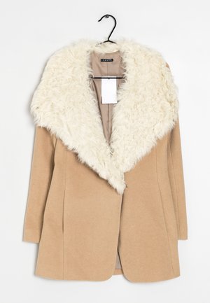 Manteau couleur camel avec grand col en fausse fourrure couleur crème suspendu sur un cintre noir sur fond blanc.