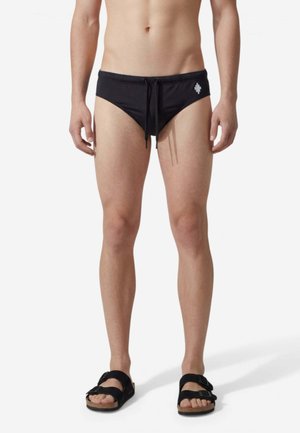 Torace e gambe maschili che indossano slip da bagno neri con logo laterale, sandali neri con fibbia, in piedi su uno sfondo bianco uniforme.