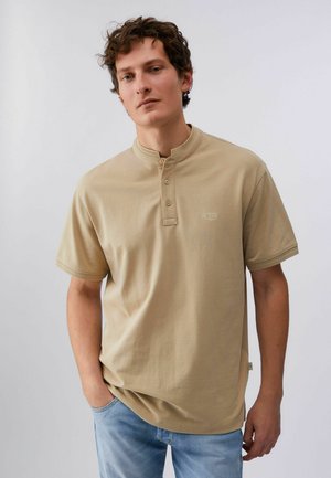 Bărbat cu păr creț, purtând o cămașă henley bej cu mâneci scurte și blugi albaștri deschiși, stând cu o mână în buzunar.