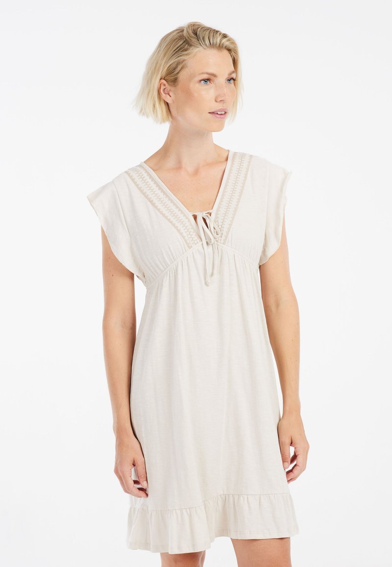 Vestido beige claro con mangas al hombro, escote en V y dobladillo con volantes. Presenta costuras decorativas en el pecho y detalle con cordón.