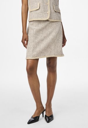 Ensemble en tricot en deux pièces de couleur crème avec un motif texturé. La jupe a une coupe droite et un ourlet jaune ; portée avec des escarpins noirs à bout pointu.