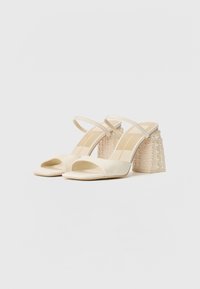 Des chaussures à talons hauts ouvertes à l'avant en tissu blanc cassé avec un talon carré orné d'accents en perles transparentes et de fines lanières aux chevilles.