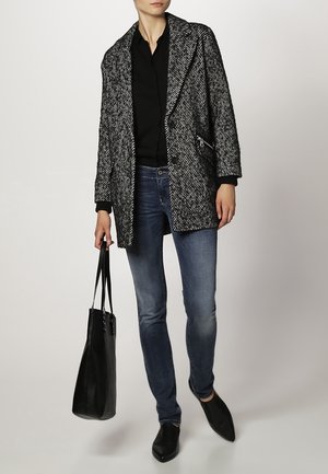 Zwart-wit houndstooth oversized jas, zwarte shirt, blauwe jeans, zwarte tote bag en zwarte schoenen met spitse neuzen.