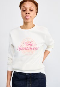 Bílá mikina se růžovým logem "Nike Sportswear", kulatý výstřih, žebrované manžety a měkká textura. Nosí se s tmavými džíny.