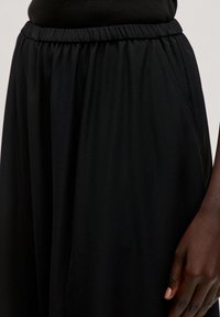 Zwarte wijde broek met een elastische tailleband. De stof is glad en iets gestructureerd, met zijzakken voor functionaliteit.