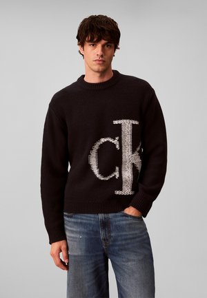 Jeune homme portant un pull noir avec de grandes lettres blanches "ck" et un jean bleu délavé, debout devant un fond gris uni.