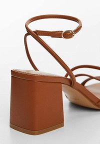 Brown heeled sandal featuring a chunky square heel, thin cross-strap design, and a circular buckle accent for adjustable fit.