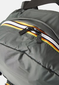 Mochila de tela gris con un bolsillo con cremallera y un acento de rayas multicolores. Las cremalleras son de plástico negro con detalle de logo.