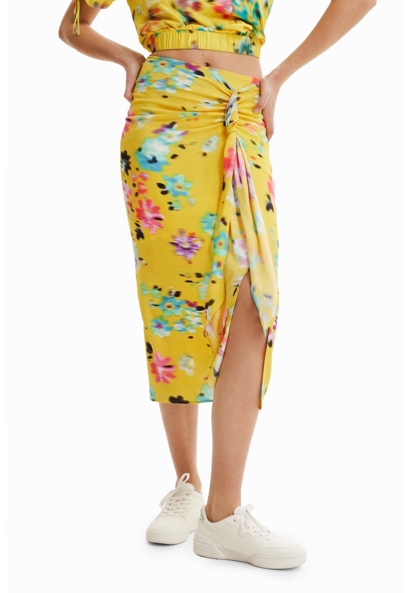 Jupe midi fleurie jaune avec un nœud sur le côté et une fente, portée avec des baskets blanches et un top court assorti.