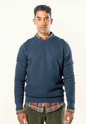 Marineblauer strukturiertes Strickpullover mit Rundhalsausschnitt, über einem mehrfarbigen karierten Hemd getragen, mit langen Ärmeln und gerippten Säumen.