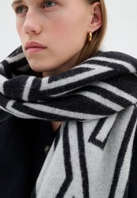 Donna con capelli corti biondi che indossa orecchini a cerchio dorati e una sciarpa a motivi geometrici bianco e nero sopra un cappotto scuro.