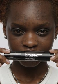 Fondotinta MAC Studio Fix Every-Wear, in una bottiglia cilindrica nera, tenuta da una persona con pelle strutturata e capelli ricci, contro uno sfondo neutro.