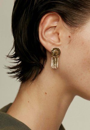 Pendientes de oro con una base circular que presenta una superficie texturizada y un diseño central, adornados con múltiples flecos metálicos colgantes.