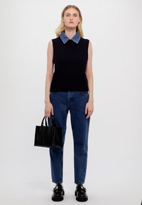 Marinekleurige mouwloze sweater met een denim kraag, gecombineerd met blauwe jeans en een zwarte handtas. Model draagt glanzende zwarte schoenen. Eenvoudig, modern ontwerp.