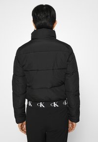 Svart puffjacka med hög krage, quiltat mönster och elastisk midja med vitt logotyp. Färgen är enhetlig utan synliga mönster.