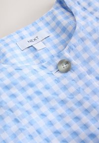 Tissu vichy bleu et blanc avec un bouton gris rond et une étiquette blanche cousue portant l’inscription « NEXT » au col.