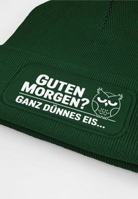 Green knitted beanie featuring a textured fabric, a rectangular patch with white text "GUTEN MORGEN? GANZ DÜNNES EIS..." and an owl graphic.