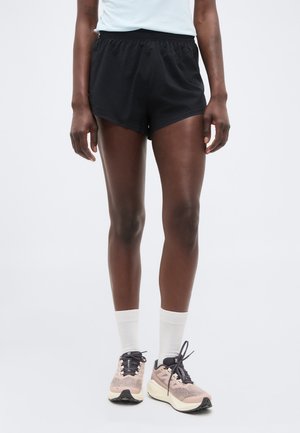 Άτομο που φοράει μαύρα αθλητικά σορτς, λευκές κάλτσες πλήρους μήκους και ροζ παπούτσια για τρέξιμο με μαύρα κορδόνια, στέκεται μπροστά σε απλό φόντο.