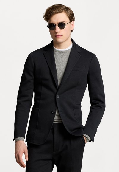 Zwarte blazer met een gladde textuur, sluiting met één knoop, twee voorzakken, gedragen over een grijs sweater. Ronde zonnebril maakt de look compleet.