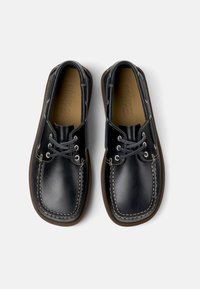 Zapatos de cuero negro con punta redonda, cordones suaves y costuras blancas que contrastan. Presentan una suela de goma marrón y un diseño sencillo.