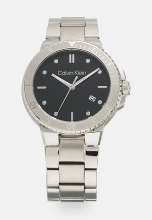 Reloj de acero inoxidable con esfera negra, manecillas plateadas y marcadores de horas. Pantalla de fecha a las 6 en punto. Pulsera de estilo industrial.
