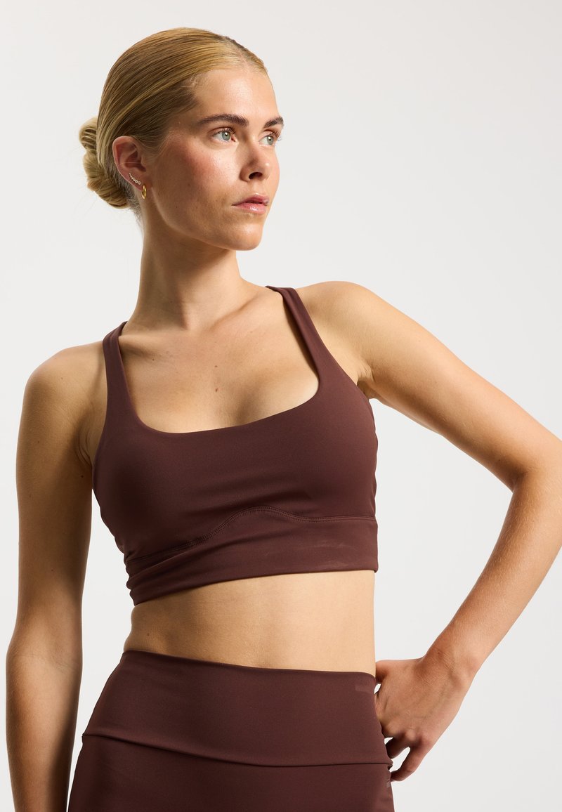 Brassière de sport marron avec un décolleté arrondi et de larges bretelles, fabriquée en tissu lisse et extensible. Elle présente un design sans coutures et une coupe ajustée.