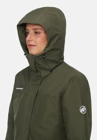Olijfgroene waterdichte jas met een capuchon, voorzien van een ritssluiting aan de voorkant, verstelbare trekkoorden en een ronde logo op de mouw.