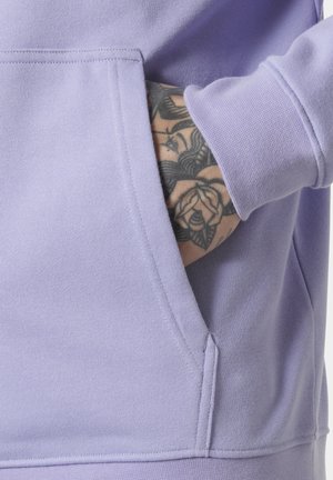 Hand met bloemtattoo in de voorkantzak van een lavendelkleurige trui met zichtbare naadstiksels.