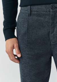 Męska ręka spoczywająca obok szarych w kratkę spodni i czarnej koszuli z długim rękawem, ukazująca fakturę materiału oraz detal guzików.