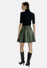 Schwarzes Kurzarmoberteil, kombiniert mit einem grünen A-Linien-Rock, der einen Reißverschluss auf der Rückseite hat. Das Modell trägt kniehohe schwarze Stiefel mit spitzem Kopf.