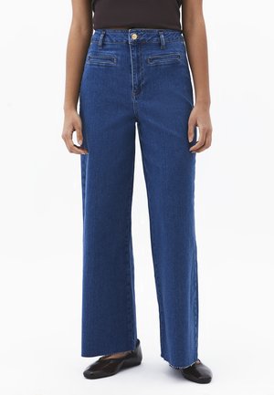 Person, der hoch taillierte, weit geschnittene blaue Jeans mit vorderen Leistentaschen und schwarzen flachen Schuhen vor einem schlichten Hintergrund trägt.