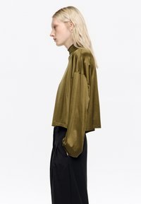 Oliwkowa bluzka z długim rękawem i wysokim dekoltem, luźny krój z opadającymi ramionami, zestawiona z czarnymi spodniami o wysokim stanie.