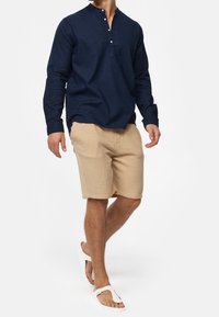 Maglia a maniche lunghe blu navy con collo henley con bottoni, abbinata a pantaloncini beige chiaro con trama e infradito bianche, su una figura in cammino.