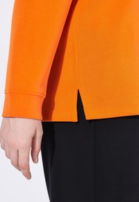 Maglietta arancione a maniche lunghe con dettaglio a fessura laterale, caratterizzata da una texture morbida e una vestibilità rilassata, abbinata a un tessuto nero sottostante.
